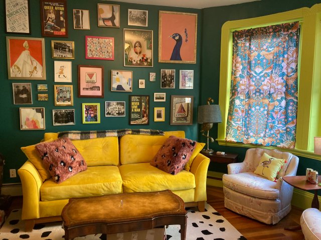 Living room with dark turquoise walls, chartreuse trim on the window frame, a bright yellow couch, a black and white dotted rug, rust-colored pillows, and a wide variety of framed prints of different sizes and colors